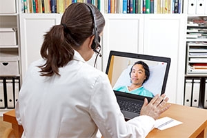 image of the back of a female teleneurohospitalist with a headset on her head gazing at a computer screen with a male patient visible onscreen